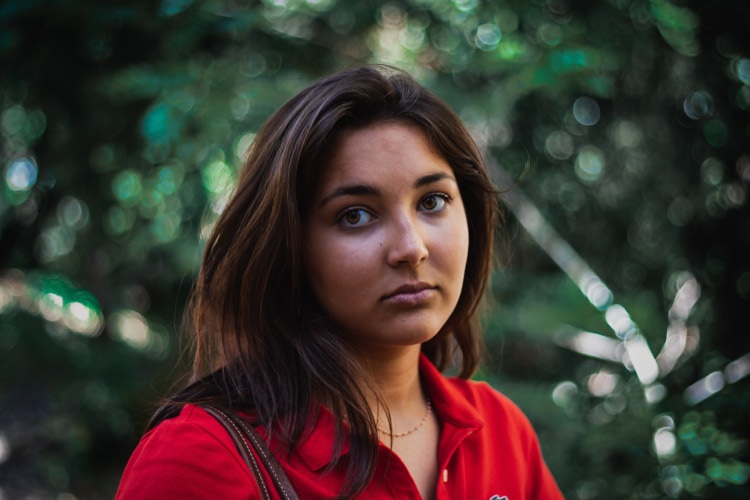 Brunette young woman with dental anxiety wears a red blouse while standing near green foliage