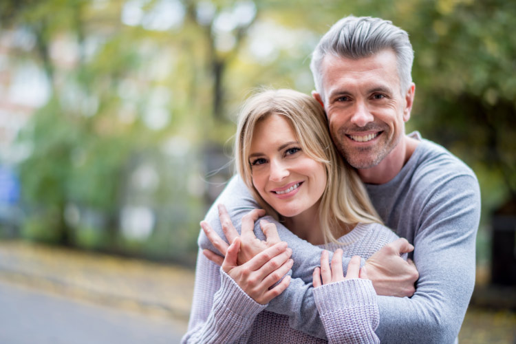 A middle-aged husband and wife embrace outside while smiling with dental implants