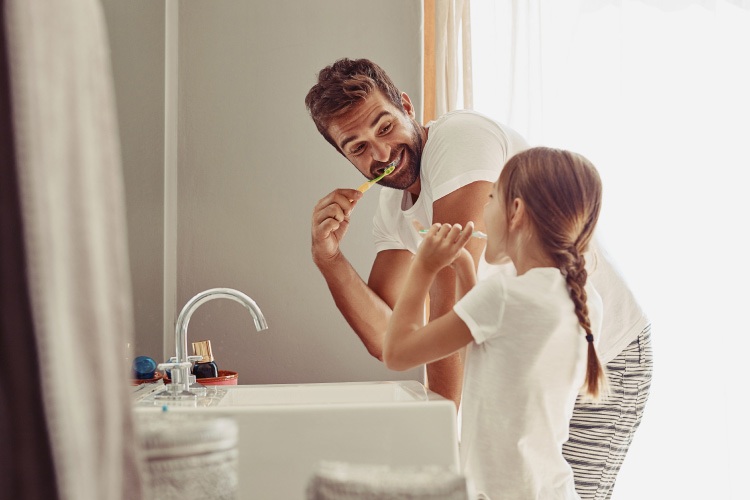 A dad brushes his teeth next to his daughter at the bathroom sink