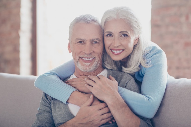 An older couple smiles with dentures and embrace while sitting on a couch