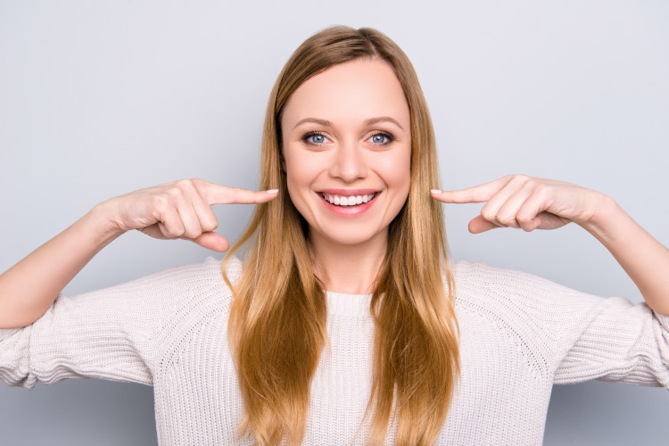 Blonde woman points to her professionally whitened smile while wearing a white sweater