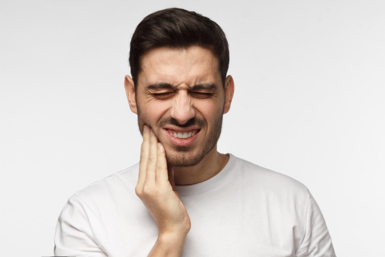 Brunette man wearing a white shirt cringes in pain due to an emergency toothache