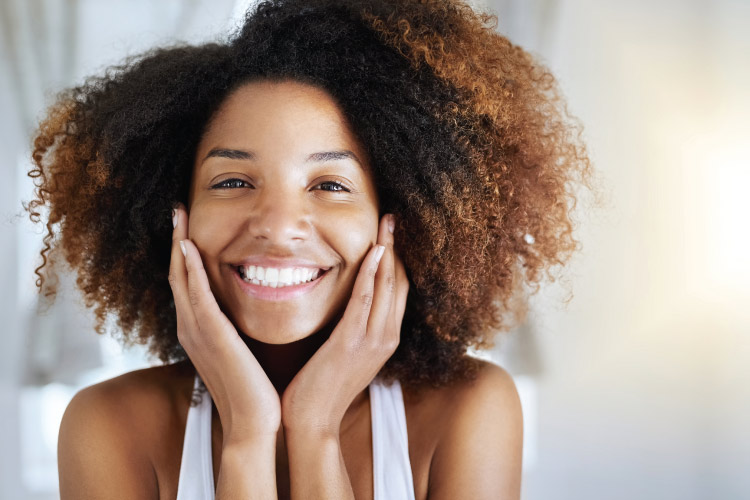 Woman with curly brown hair smiles with veneers and touches her cheeks while wearing a white tanktop