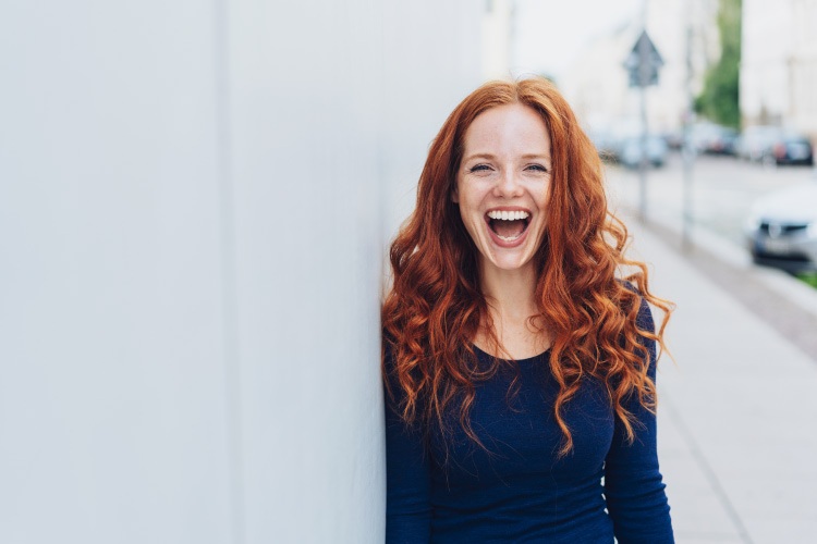 Red haired woman with porcelain veneers smiles while standing against a white wall outside