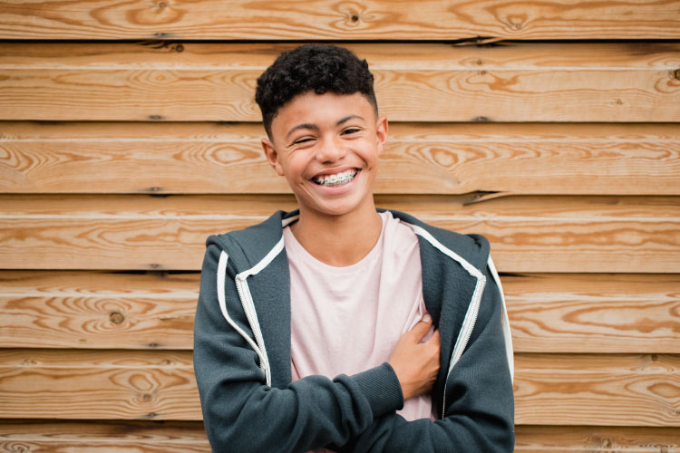Smiling brown skinned teenage boy wearing braces stands in front of a horizontal fence.