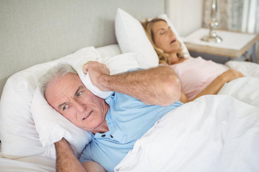 Man covering his ears with a pillow while his wife with sleep apnea snores away next to him in bed.
