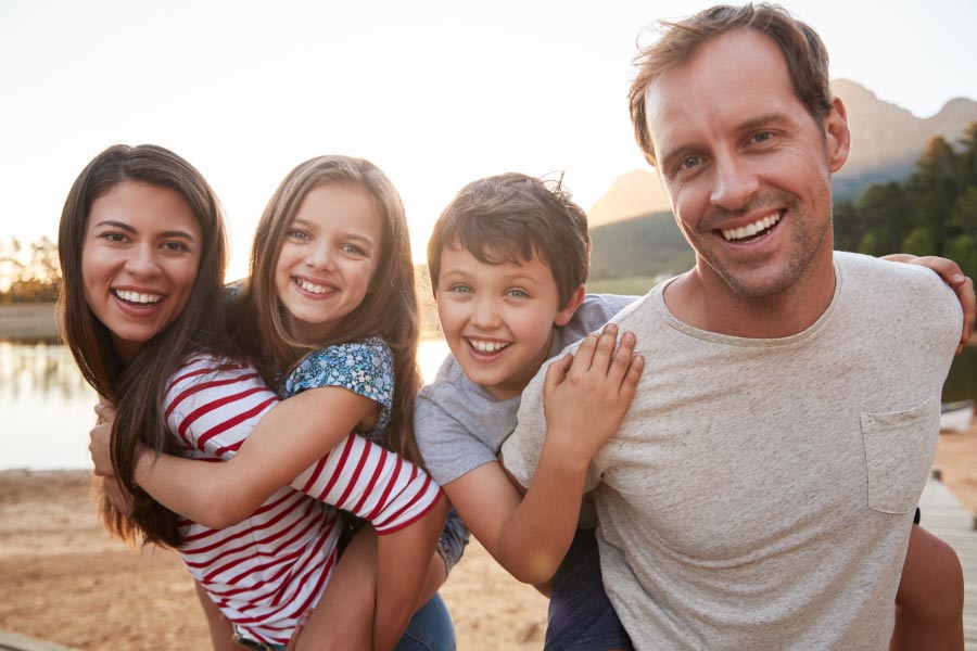 Young family near a lake with a boy on the dad's back and a girl on the mom's back.