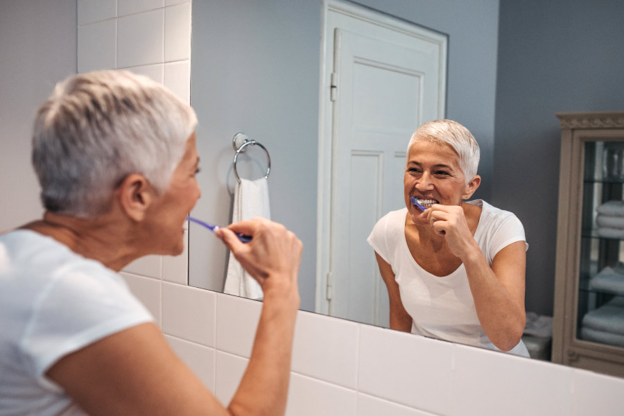 Lady with short gray hair brushing her teeth in front of the mirror.